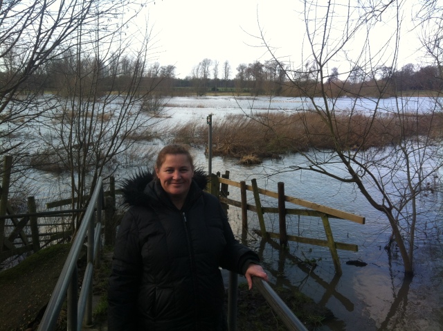 Sharlene smiles happily after finding a cache. She stands on a bridge with a view of fields in the background which are flooded due to the recent heavy rain causing the river to burst its banks.