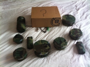 The image shows a lot of geocaches laid out. They are all covered in camoflauge tape. In the centre at the top is a box with a question mark on it covering two of the caches from view.
