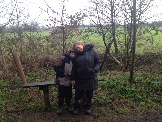 Sam and Shar sit on a bench with fields in the background. They are wrapped up against the cold.