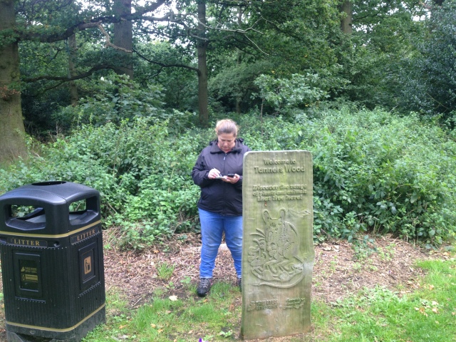 Shar stands behind a wood sculpture of a mouse that holds the sign for Tanner's wood. She is collecting numbers off the information board behind for a multi cache