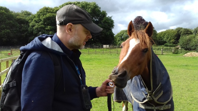 Paul stands in a field next to a horse holding his Jimmy Talon TB. The horse is just about to nibble the TB and possibly Paul's fingers.
